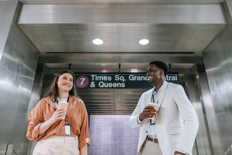 Two colleagues laugh and hold coffee near a subway sign in an underground station.